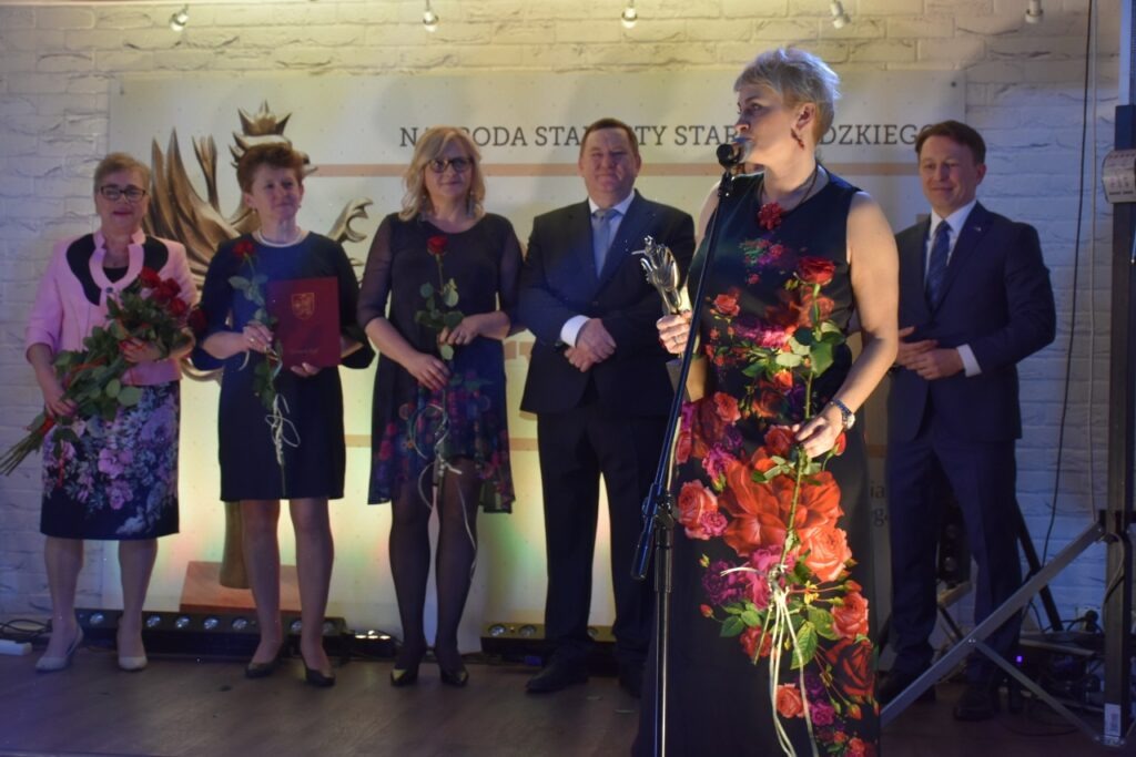 Reprezentacja zarządu powiatu i osoby nagrodzone stojące na scenie, jedna osoba mówi przez mikrofon